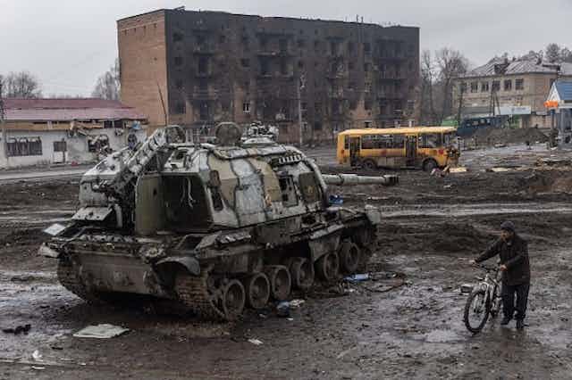 Un homme pousse son vélo dans la boue et les débris à côté d'un char russe détruit devant la gare centrale qui servait de base russe, le 30 mars 2022 à Trostyanets, en Ukraine.