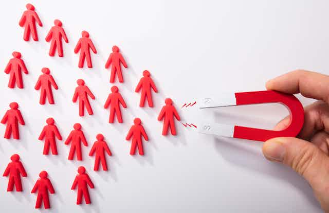 A hand holding a magnet with the ends facing toward a pyramid of red human figures. A small electric bolt is drawn between the magnet and the nearest figure.
