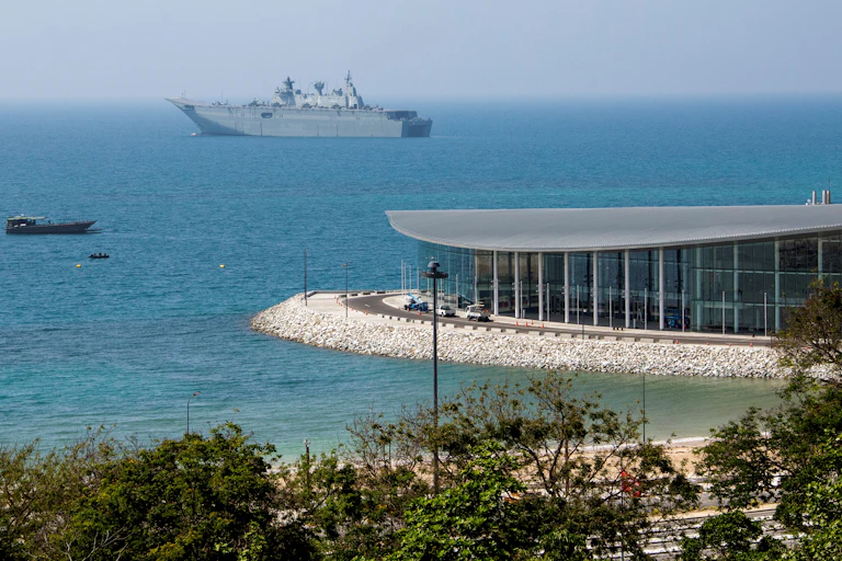 A large grey ship is seen in the distance on very blue waters, with a glass building pictured on the shore.