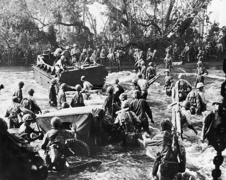 Men wearing military uniforms and helmets stand on boats and walk through the water in a black and white photo, heading toward a wild looking shore.