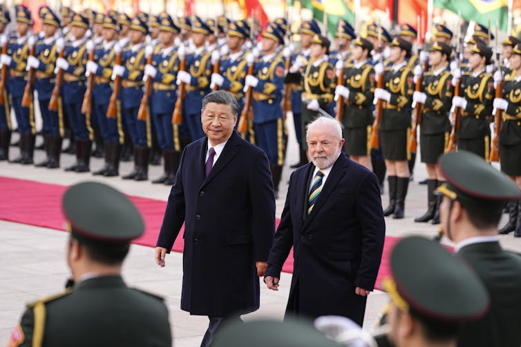 Two men in coats walk side by side in front of a parade of military men.