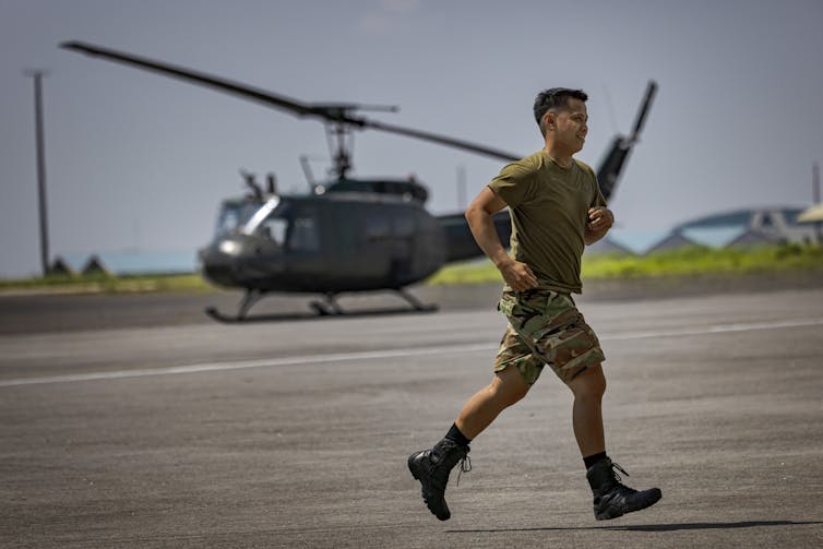 A man dressed in green shirt and shorts runs across tarmac with a helicopter visible in the background.
