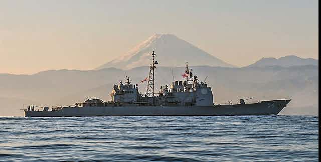 A large naval ship on the ocean with snow-capped mountain in the background.