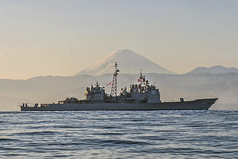 A large naval ship on the ocean with snow-capped mountain in the background.