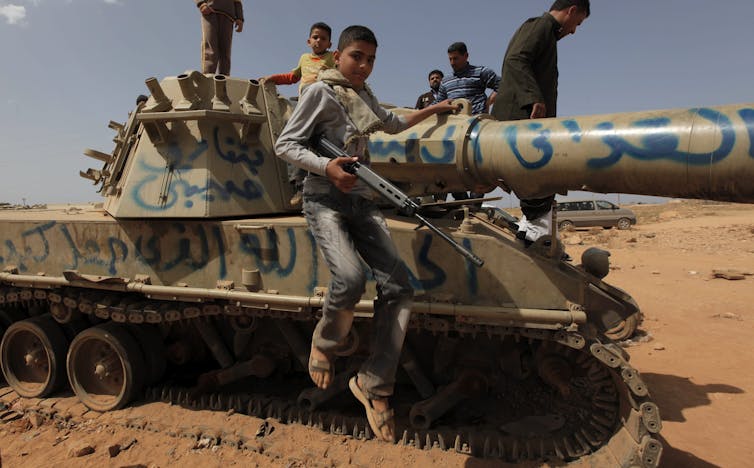 A teenaged boy carries a gun as he jumps from a tank.