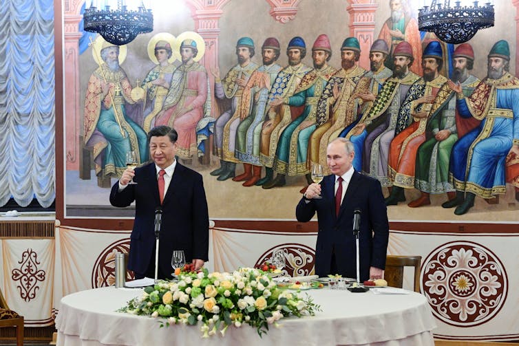 An Asian man and a balding man raise their champagne glasses with a colourful mural behind them and a large bouquet of flowers on the table in front of them.
