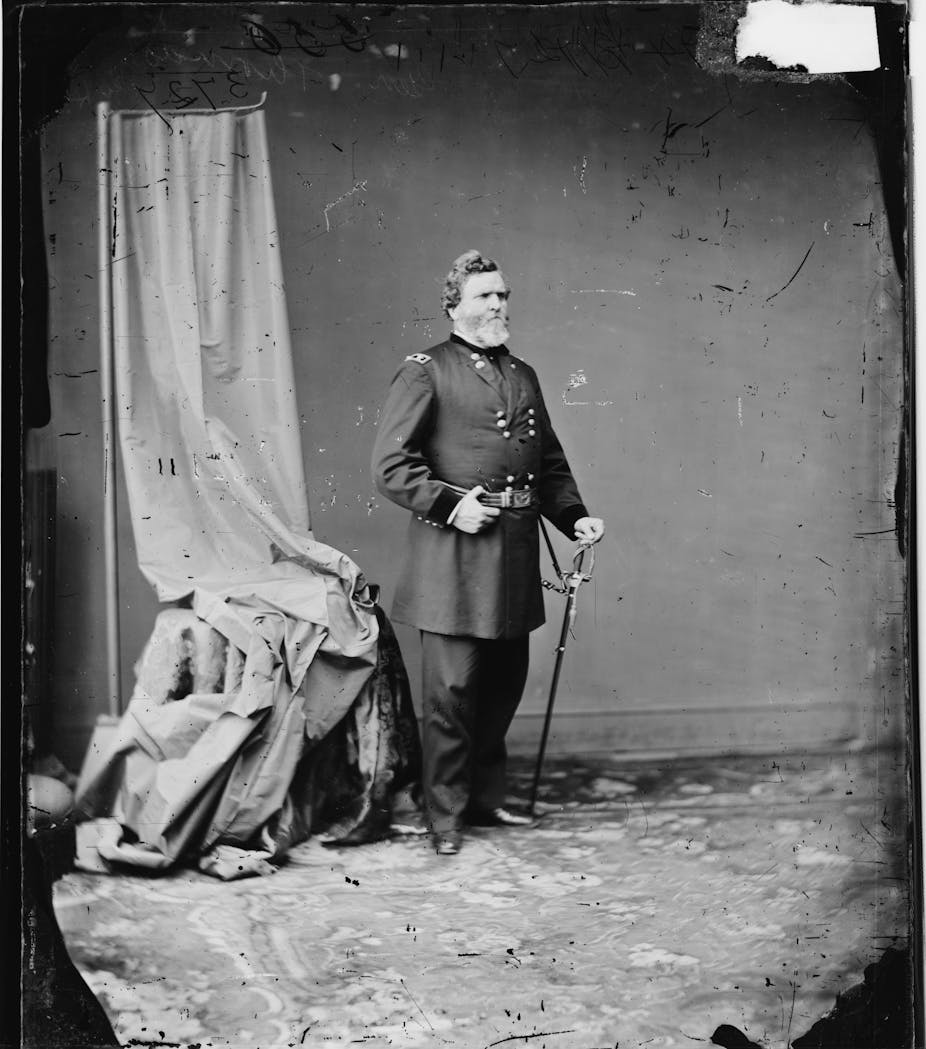 A man wearing a Union army uniform stands with a sword pointed into the ground in his left hand and clasping the belt of his uniform with his right hand. A portrait curtain hangs behind him.