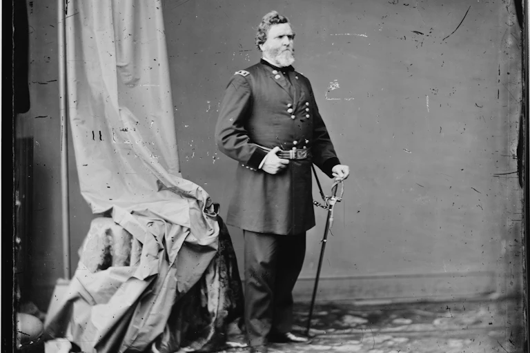 A man wearing a Union army uniform stands with a sword pointed into the ground in his left hand and clasping the belt of his uniform with his right hand. A portrait curtain hangs behind him.
