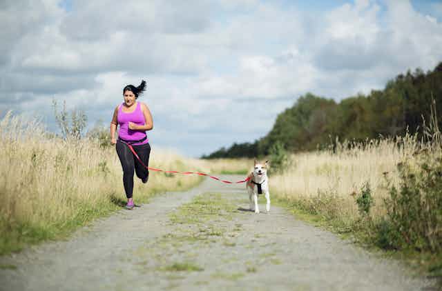 Overweight woman running on a dirt path with dog attached to her with a leash.