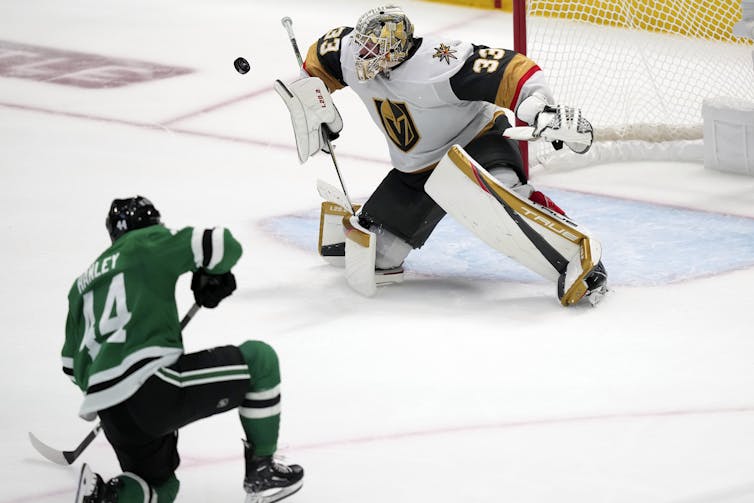 A man in a green hockey jersey shoots a puck toward a goaltender in a white jersey.