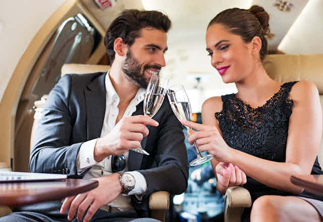 a white man and white woman wearing fancy clothes chink champagne glasses in a private jet