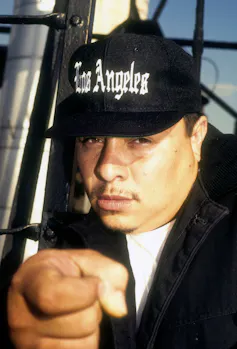 A man with a faint mustache wearing a Los Angeles baseball cap points at the camera close-up.