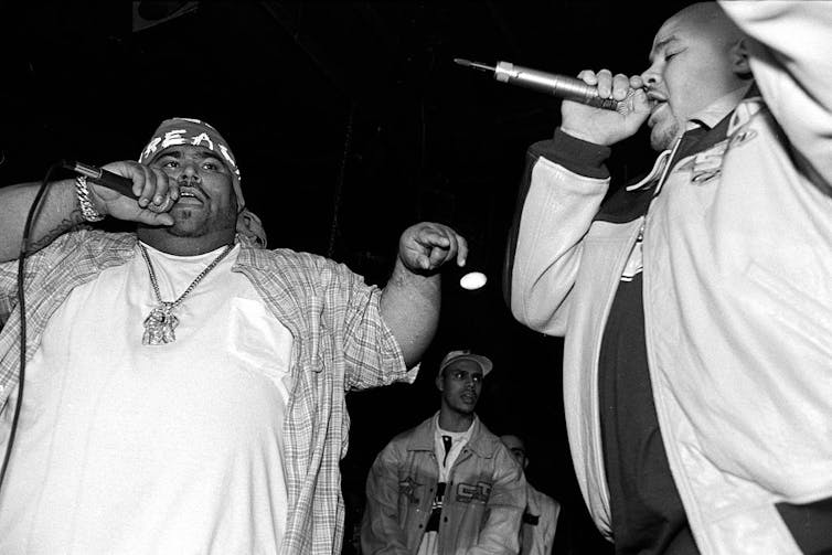 A black and white picture, taken from below a stage, of two large men rapping into microphones.