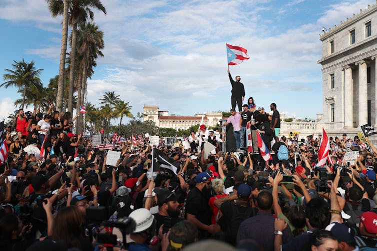 A large crowd outside, with men on a truck holding Puerto Rican flags.