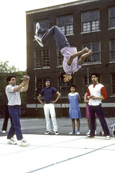 Teens stand in a schoolyard as a young man does a high back flip.