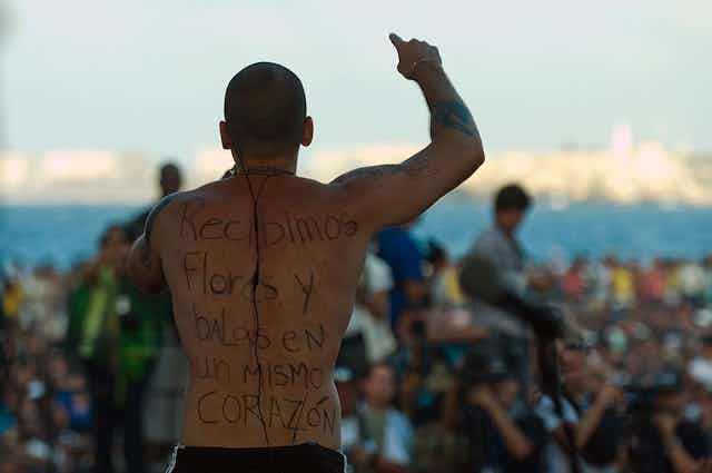 A shirtless man holds up his arms as he sings to a crowd. Written on his back is 'Recibimos flores y balas en un mismo corazon.'