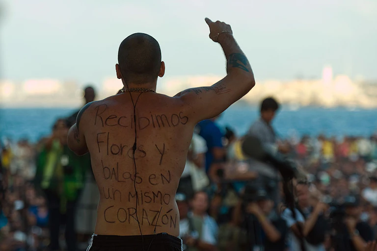 A shirtless man holds up his arms as he sings to a crowd. Written on his back is 'Recibimos flores y balas en un mismo corazon.'