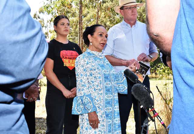Linda Burney wears a blue dress and speaks in front of microphones. Behind her stands a person who wears a t shirt supporting the Uluru Statement.