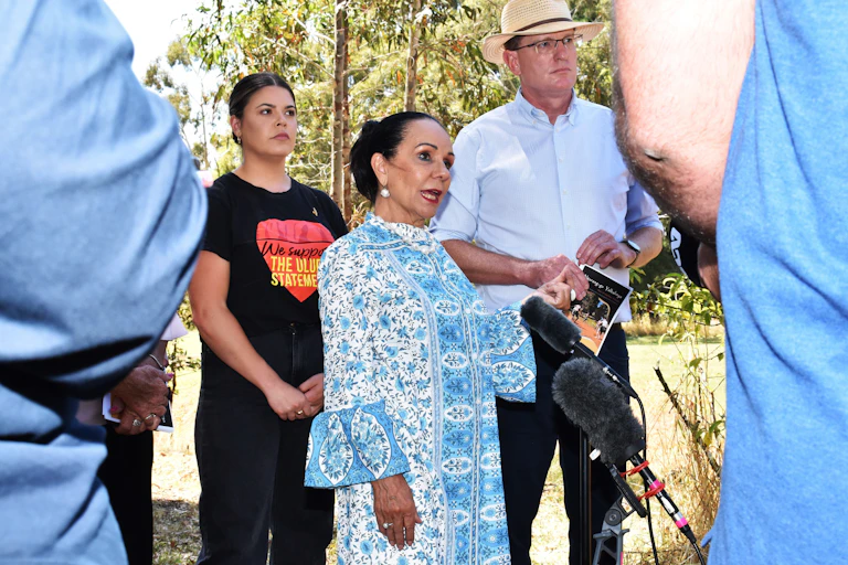 Linda Burney wears a blue dress and speaks in front of microphones. Behind her stands a person who wears a t shirt supporting the Uluru Statement.