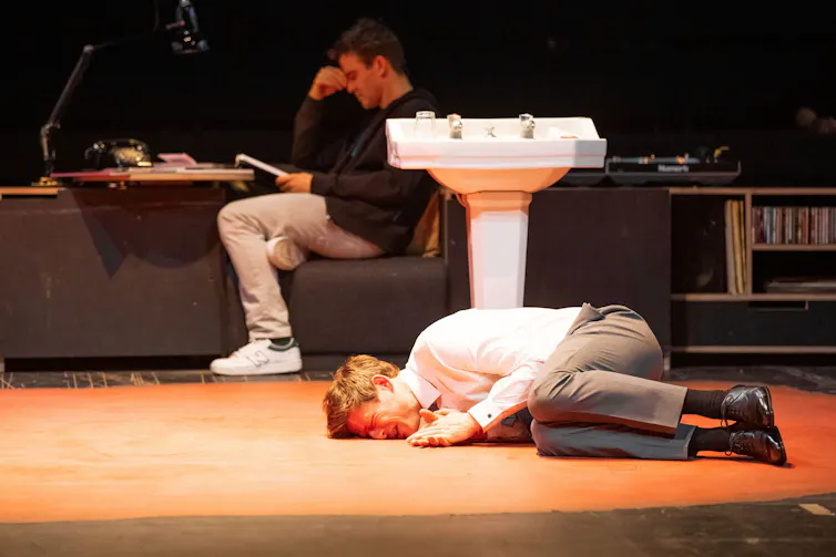 A man on stage lies in the fetal position on the floor.