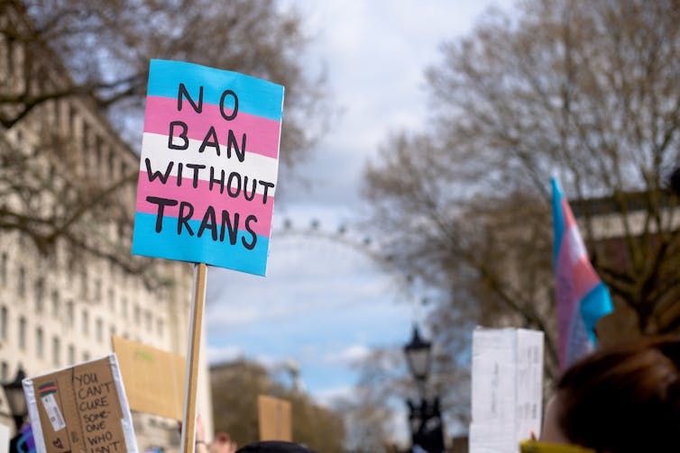 A protest sign painted in the pink, blue and white stripes of the trans pride flag, reading 'No ban without trans'.