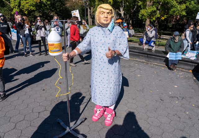 Person dressed in hospital gown wearing a Donald Trump mask.