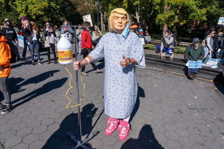Person dressed in hospital gown wearing a Donald Trump mask.