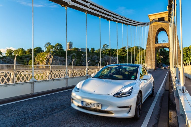 White Tesla car driving on a suspension bridge at sunset, trees and buildings in the background.