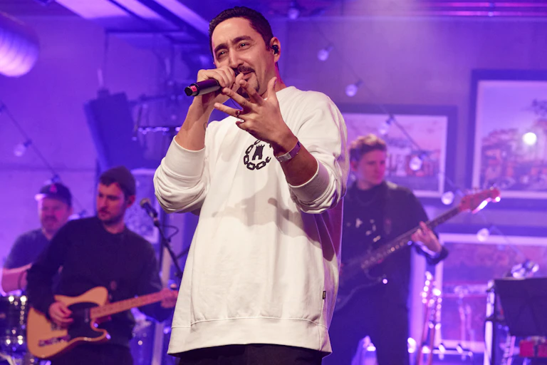 A man dressed in a long-sleeved white t-shirt is holding a microphone in front of a band.