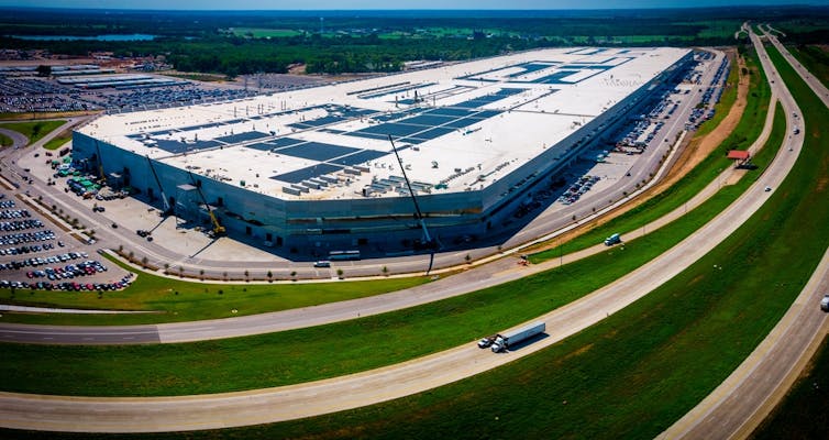 Aerial view of very large building surrounded by grass and roads, trucks, cars.