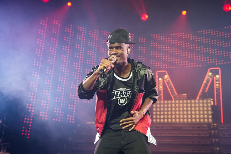 A Black man wearing a baseball cap and leather jacket holds a microphone as he sings on stage