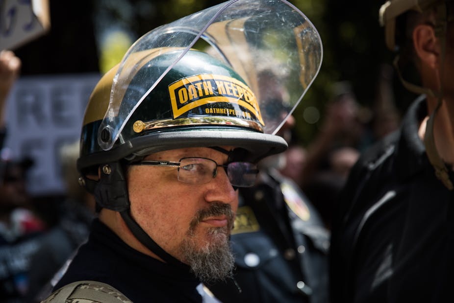 A man in a protective helmet labeled Oath Keepers stands in a crowd.
