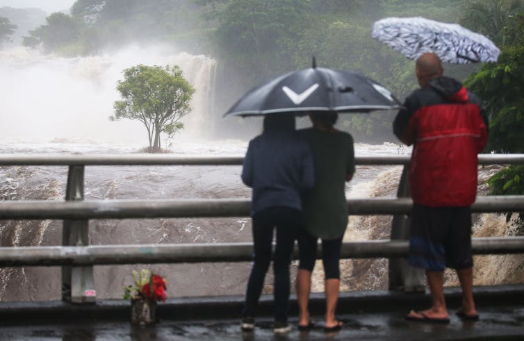 Three people stand under umbrellas on a bridge watching a rushing river below. It's clearly well beyond its banks, with a tree in the middle of the water, and moving so fast spray is coming up.
