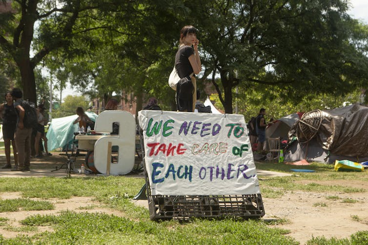 A woman in a park stands next to a sign that reads We Need to Take Care of Each Other.