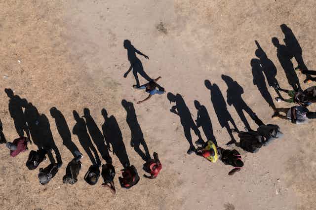 A row of people are seen waiting on dirt, as seen from an aerial view, with their shadows elongated. 