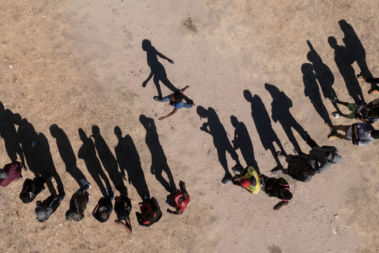 A row of people are seen waiting on dirt, as seen from an aerial view, with their shadows elongated.