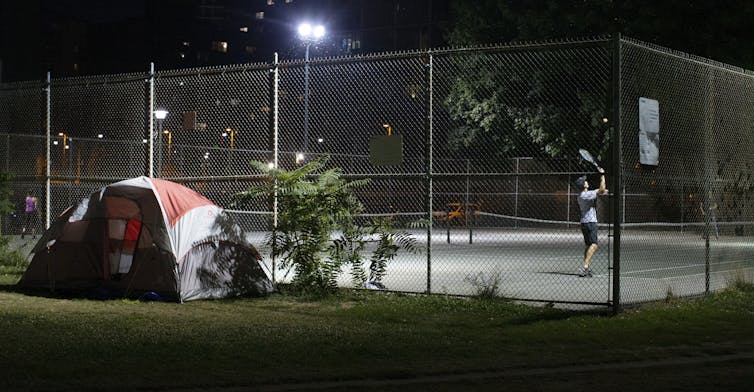 A man plays tennis on a public court with a homeless person's tent next to it.