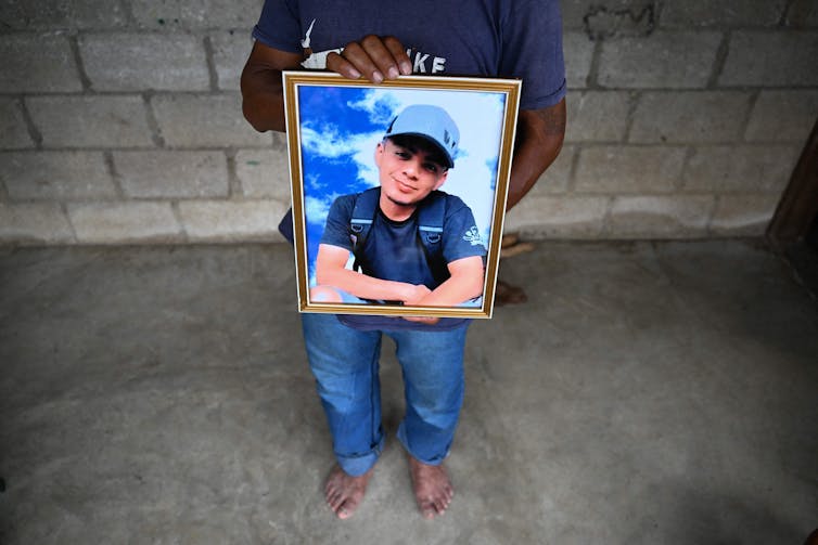 A person, seen from below the neck, holds a large framed photo of a young man, smiling, wearing a blue shirt and hat.