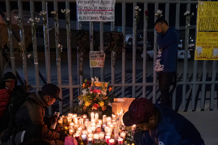 People crouch near a series of candles and photos outside of a large fence.