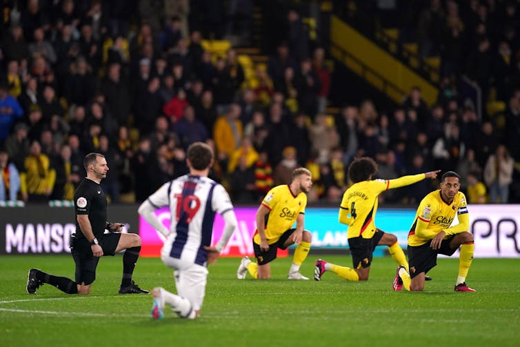 Soccer players in yellow and blue and white, and a referee in black, all kneel on the grass.