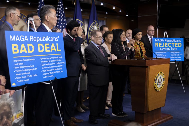 Many people in business clothing on a stage with signs that say 'MAGA Republicans' BAD DEAL.'