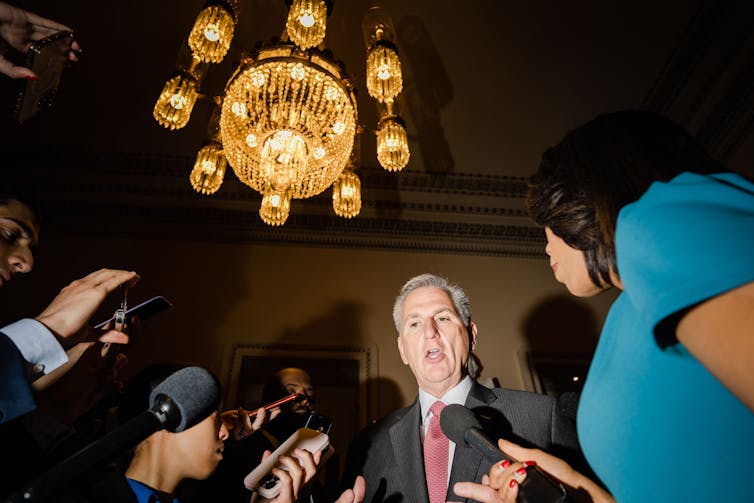 A gray haired man in suit and tie talking to reporters under a chandelier.