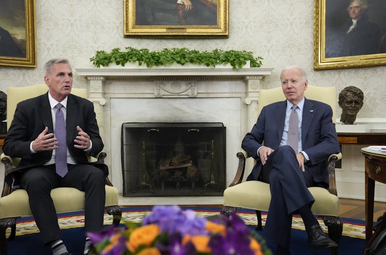 Two men sitting in yellow armchairs in front of an elegant fireplace.