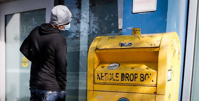 A man wearing a mask stands next to a yellow needle drop box.