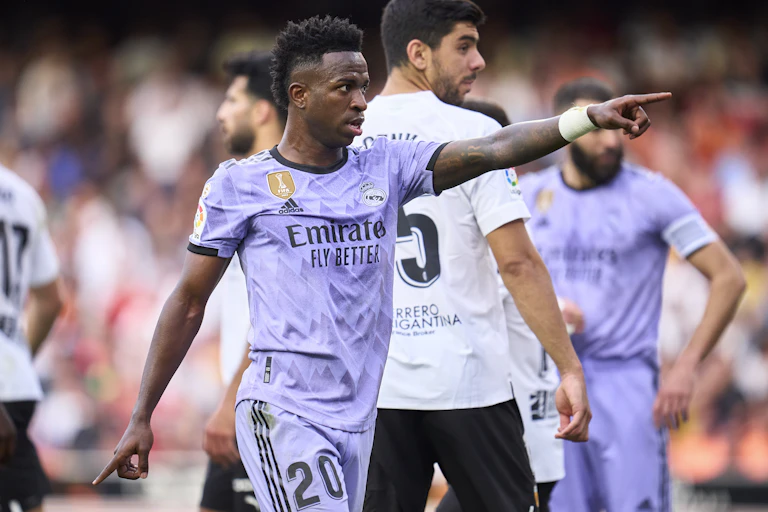 A Black man in a lilac soccer jersey points at fans in a crowd.