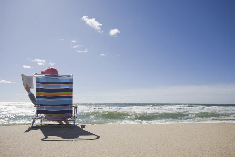 A person sits on a rainbow colored lawn chair as waves crash on the beach under a sunlit sky.