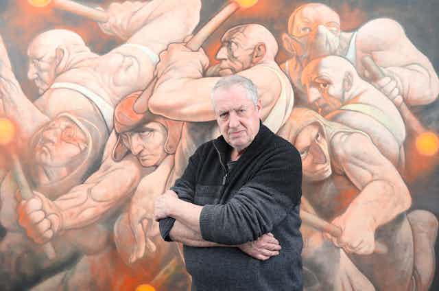 Th artist Peter Howson standing in one of his paintings showing lots of bald, aggressive-looking men in white vests.