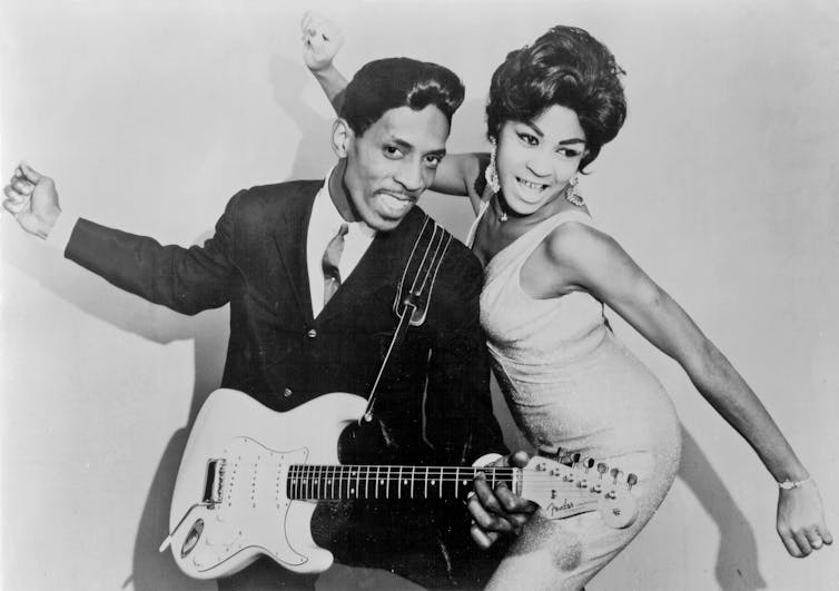 A young Black couple in a black and white photograph with the man holding an electric guitar.