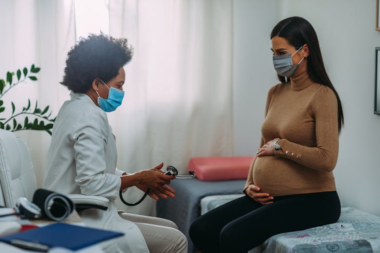 A pregnant woman and a doctor both wearing face masks in the doctor's office