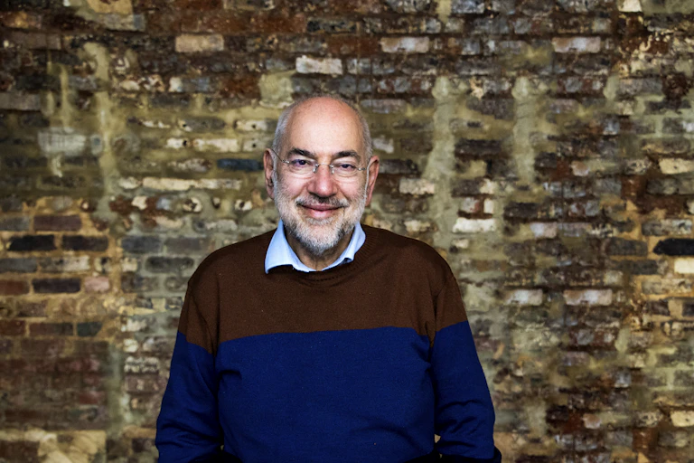 A smiling, bespectacled man in a collared shirt beneath a two-toned jumper stands in front of a brick wall
